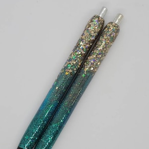 May include: Two teal and gold glitter pens with silver accents. The pens have a gradient of glitter, starting with gold at the top and fading to teal at the bottom.