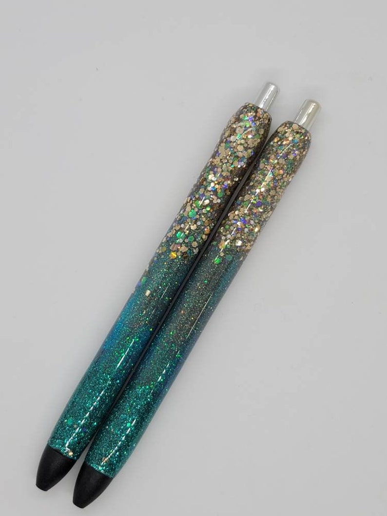 May include: Two teal and gold glitter pens with silver accents. The pens have a gradient of glitter, starting with gold at the top and fading to teal at the bottom.