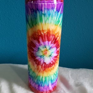 May include: A stainless steel tumbler with a black lid and a colorful tie-dye design. The design features a swirling pattern of red, orange, yellow, green, blue, and purple.