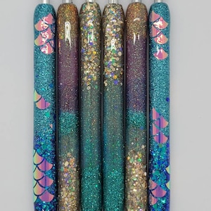 May include: Six glitter pens with iridescent mermaid scales and a black ink tip. The pens are filled with different colors of glitter, including gold, teal, and purple.