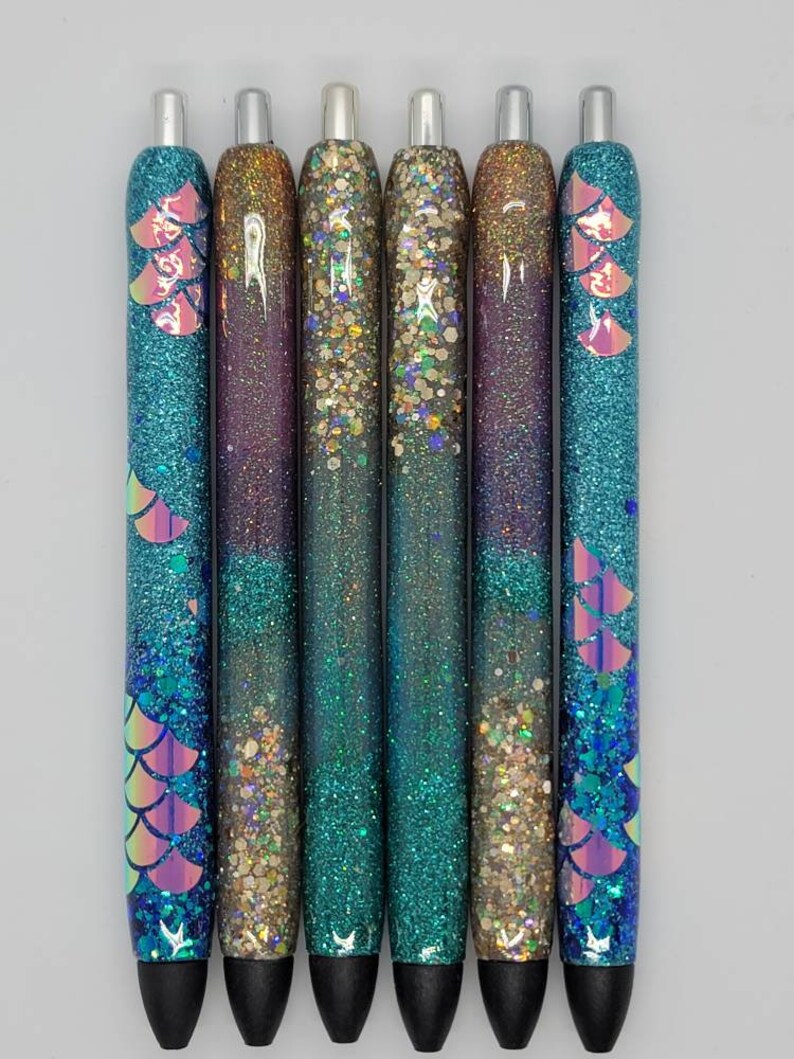 May include: Six glitter pens with iridescent mermaid scales and a black ink tip. The pens are filled with different colors of glitter, including gold, teal, and purple.