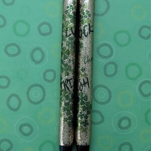 May include: Two gold glitter pens with black ink and black caps. The pens have green glitter and the words "Good Luck" and "Irish" written on them.