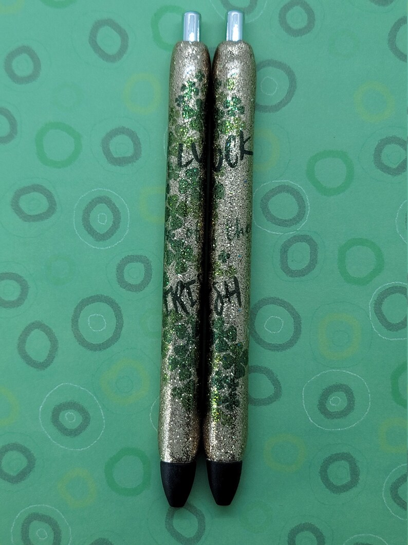May include: Two gold glitter pens with black ink and black caps. The pens have green glitter and the words "Good Luck" and "Irish" written on them.