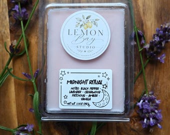 Könnte beinhalten: Ein hellrosa Duftwachs in einer durchsichtigen Kunststoffverpackung. Das Etikett zeigt den Text Lemon Bay Studio und ein zweites Etikett trägt die Aufschrift Midnight Ritual mit Noten von schwarzem Pfeffer, Lavendel, Zedernholz, Patchouli, Amber und Vanille, 71g.