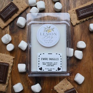May include: A rectangular plastic clamshell container filled with white wax melts. The top label features a lemon illustration and the text Lemon Bay Studio. The bottom label reads S'MORE SNUGGLES, notes of warm spice, velvet woods, sugared vanilla, golden amber, and net wt. 2.5 oz (71g).
