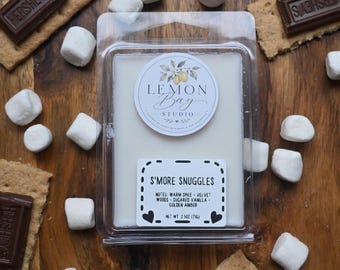 Könnte beinhalten: Ein rechteckiger Kunststoffbehälter gefüllt mit weißem Wachs. Das obere Etikett zeigt eine Zitronenillustration und den Text Lemon Bay Studio. Das untere Etikett trägt die Aufschrift S'MORE SNUGGLES, notes of warm spice, velvet woods, sugared vanilla, golden amber, und net wt. 2.5 oz (71g).