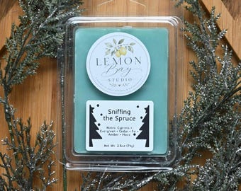 Könnte beinhalten: Ein rechteckiges, salbeigrünes Wachsschmelzstück in einer durchsichtigen Kunststoffverpackung. Das Etikett zeigt den Text Lemon Bay Studio und Sniffing the Spruce mit Duftnoten von Zypresse, immergrün, Zeder, Tanne, Amber und Moos. Nettogewicht 71g.