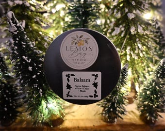 Könnte beinhalten: Eine runde schwarze Kerzendose mit einem Lemon Bay Studio Logo-Aufkleber und einem weißen rechteckigen Etikett mit der Aufschrift Balsam, Notes: Balsam, Pine, Cedar, Musk, Net Wt. 7oz (198g), vor mehreren kleinen künstlichen Tannen.
