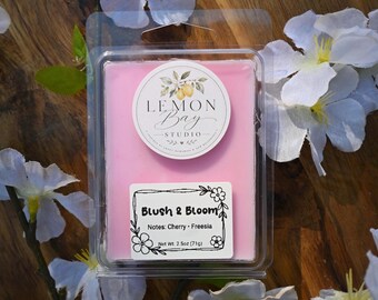 Könnte beinhalten: Ein rechteckiges rosa Duftwachs in einer durchsichtigen Kunststoffverpackung. Das obere Etikett zeigt den Text Lemon Bay Studio mit einer Zitronenillustration. Ein unteres Etikett trägt die Aufschrift Blush & Bloom, Notes: Cherry - Freesia, 71g.
