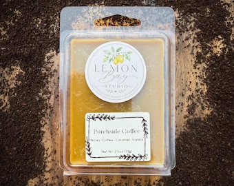 Könnte beinhalten: Ein rechteckiges Duftwachs in einer durchsichtigen Kunststoffverpackung. Das Wachs ist goldbraun. Ein rundes Etikett zeigt Lemon Bay Studio mit einer Zitronenillustration. Ein rechteckiges Etikett trägt die Aufschrift Porchside Coffee, Notes Coffee, Caramel, Vanilla, Net Wt. 71g.