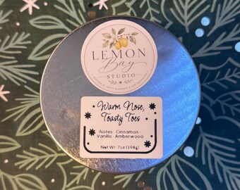 Könnte beinhalten: Eine runde Kerzendose aus silbernem Metall mit zwei Etiketten. Das obere runde Etikett zeigt eine Zitronenillustration und den Text Lemon Bay Studio. Das rechteckige Etikett trägt die Aufschrift Warm Nose, Toasty Toes, Notes Cinnamon, Vanilla, Amberwood, Net Wt 7oz (198g).