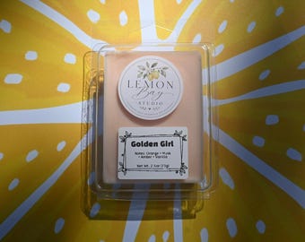 Könnte beinhalten: Ein rechteckiges, pfirsichfarbenes Duftwachs in einer durchsichtigen Kunststoffverpackung. Das obere Etikett zeigt Lemon Bay Studio mit einer Zitronenillustration. Das untere Etikett trägt die Aufschrift Golden Girl, Notes: Orange, Musk, Amber, Vanilla, und Net Wt. 2.5oz (71g).