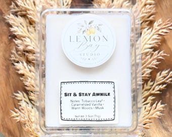 Könnte beinhalten: Eine rechteckige, durchsichtige Kunststoffverpackung mit einem weißen Duftwachs-Tab. Das obere Etikett zeigt den Text Lemon Bay Studio mit einer Zitronenillustration. Das untere Etikett trägt die Aufschrift Sit & Stay Awhile mit Noten von Tabakblatt, karamellisierter Vanille, warmen Hölzern, Moschus und einem Nettogewicht von 71g.