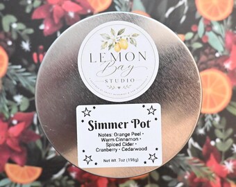 Könnte beinhalten: Ein runder silberner Metalldosendeckel mit einem kreisförmigen Lemon Bay Studio Logo und einem rechteckigen weißen Etikett mit schwarzem Text: Simmer Pot, Notes: Orange Peel, Warm Cinnamon, Spiced Cider, Cranberry, Cedarwood, 198g.