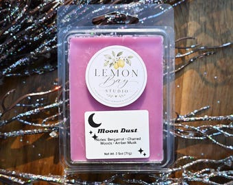 Könnte beinhalten: Ein rechteckiges violettes Duftwachs in einer durchsichtigen Kunststoffverpackung. Das Etikett zeigt das Lemon Bay Studio Logo und einen weißen Aufkleber mit dem Text Moon Dust, Noten von Bergamotte, verbranntem Holz, Amber-Moschus und 71g.