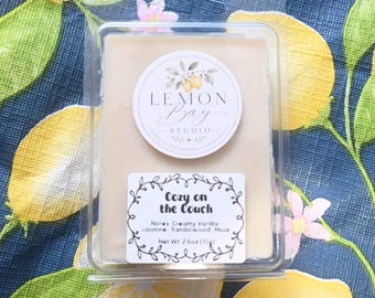 Könnte beinhalten: Eine rechteckige Duftwachspackung in einem durchsichtigen Kunststoffbehälter. Das obere Etikett zeigt den Text Lemon Bay Studio mit einer Zitronenillustration. Das untere Etikett trägt die Aufschrift Cozy on the Couch, Notes: 71g.
