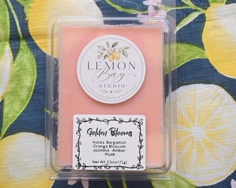 Könnte beinhalten: Ein rechteckiges, pfirsichfarbenes Wachsschmelzstück in einer durchsichtigen Kunststoffverpackung. Das Etikett zeigt den Text Lemon Bay Studio und ein zweites Etikett trägt die Aufschrift Golden Blooms, Notes Bergamot, Orange Blossom, Jasmine, Amber, Musk, 71g.