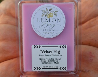 Könnte beinhalten: Ein rechteckiges rosa Wachsschmelzstück in einer durchsichtigen Kunststoffverpackung. Das obere Etikett zeigt eine Zitronenillustration und den Text Lemon Bay Studio. Ein unteres Etikett nennt Velvet Fig, Brown Sugar & Fig Inspired, Noten und 71g.