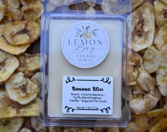 Könnte beinhalten: Ein rechteckiger Wachsschmelzbehälter aus klarem Kunststoff mit einem Block aus cremefarbenem Wachs. Das obere Etikett zeigt Lemon Bay Studio, das untere Etikett Banana Bliss mit Noten von cremiger Banane, Marshmallow, Vanille und gezuckertem Tortenboden, Nettogewicht 71g.