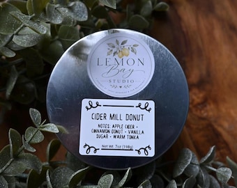 Könnte beinhalten: Eine runde Kerzendose aus silbernem Metall mit zwei Etiketten. Das obere Etikett zeigt eine Zitronenillustration und den Text Lemon Bay Studio. Das untere Etikett lautet Cider Mill Donut, Notes: Apple Cider, Cinnamon Donut, Vanilla Sugar, Warm Tonka, Net Wt. 7oz (198g).