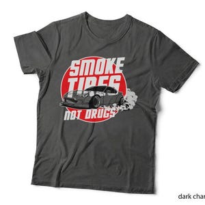 May include: A dark charcoal grey t-shirt featuring a graphic of a sports car drifting with tyre smoke. The design includes a red circle background and the white text SMOKE TIRES NOT DRUGS. The shirt has short sleeves and a crew neckline.
