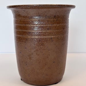 May include: A cylindrical brown ceramic vessel with a speckled, textured finish and a wide, flared rim. The exterior surface features three horizontal incised lines encircling the upper portion of the body, creating a subtle decorative detail.