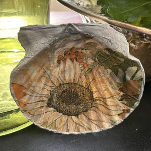 May include: A scallop shell featuring a decoupage sunflower design with orange berries and green leaves. The shell has a painted silver edge and a glossy finish, showcasing a detailed floral pattern on its curved, textured surface.