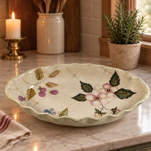 May include: A round, cream-colored ceramic serving bowl with a scalloped edge. The interior features a hand-painted pattern of pink flowers, green leaves, and clusters of purple blackberries set against a faint, light-colored grid design.