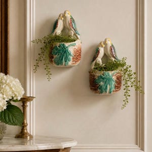 May include: Two ceramic wall planters shaped like nests, each featuring a pair of birds perched on the rim. The planters have a textured brown base with a prominent green leaf design and contain small green trailing plants.