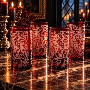May include: A set of four cylindrical glass tumblers in a deep red hue, each featuring an intricate, etched white scrollwork pattern that wraps around the exterior surface. The glasses are arranged in a cluster, displaying a consistent design.