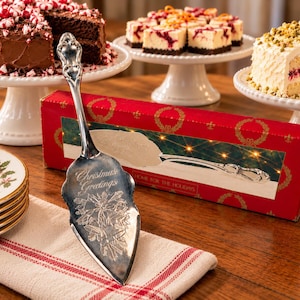 May include: A silver-toned metal cake server featuring an ornate handle and an engraved blade with the text Christmas Greetings above a floral design. The server is positioned in front of its red rectangular packaging box.
