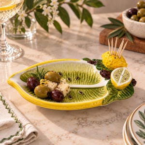 May include: A ceramic serving dish shaped like a lemon slice with a textured green interior and yellow rim. The dish features a three-dimensional yellow lemon accent on one side that serves as a holder for several wooden toothpicks.