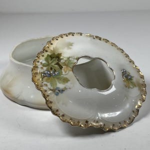 May include: A white porcelain trinket box with a removable lid featuring a central circular opening. The lid is decorated with hand-painted green leaves, small blue berries, and a scalloped gold-trimmed edge. The base is a matching white bowl.
