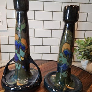 May include: Two dark ceramic candle holders with a glossy finish, featuring a tapered neck and a wide circular base. The surface is decorated with abstract painted patterns in shades of blue, green, and orange against a dark background.