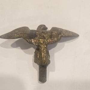 May include: A metallic, brass-toned eagle figurine with wings spread wide in a horizontal position. The eagle is mounted on a short, threaded metal bolt extending from its base, designed for attachment. The surface has a textured, aged finish.