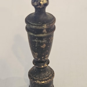 May include: A small, vertical finial with a distressed black finish and visible gold-toned wear along the edges and surface. The object features a cylindrical base, a tapered midsection, and a rounded, knob-like top, showing a weathered texture.