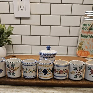 May include: A set of seven small ceramic cups and one lidded container, each featuring hand-painted floral and vine motifs in blue, yellow, green, and red on a white background with decorative scalloped edges along the rims and bases.