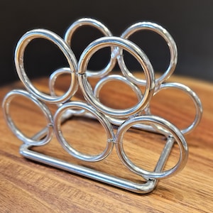 May include: A silver-toned metal napkin holder constructed from a series of interconnected circular rings welded together. The structure features a rectangular base frame that supports the vertical arrangement of multiple open metal loops.