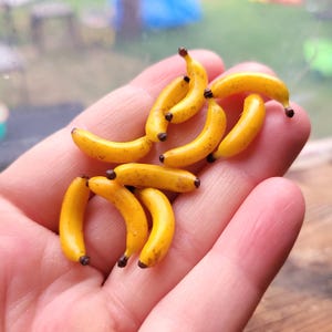 May include: A collection of small, yellow, curved banana-shaped charms with brown tips. Each miniature fruit features a smooth, glossy finish and realistic detailing, showing subtle brown speckles across the surface of the yellow peels.