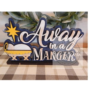 May include: Wooden Christmas decoration with the words "Away in a Manger" in white script over a navy blue background. The decoration includes a yellow star and a manger scene.