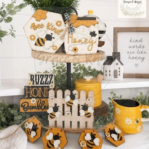 May include: A tiered display of bee-themed decor. Includes a wooden sign that says "Bee Kind", a jar that says "Honey", and a banner with bee and flower cutouts. The color scheme is yellow, black, and white.