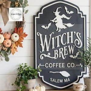 May include: A dark gray, shield-shaped wooden sign with white raised lettering that reads Witch's Brew Coffee Co. Salem MA. The sign features a white cutout of a witch on a broomstick, three bats, and a broom icon. It has a white border trim.