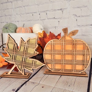 May include: Two wooden fall decorations, a pumpkin and a maple leaf, both with a plaid pattern. The pumpkin is orange and the leaf is green.