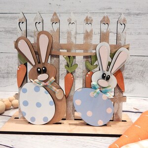Pu&ograve; includere: Decorazione pasquale in legno con due coniglietti che tengono uova a pois, davanti a una staccionata con carote. I coniglietti sono marroni e bianchi con orecchie rosa, e le uova sono blu e bianche. Un'affascinante esposizione primaverile.