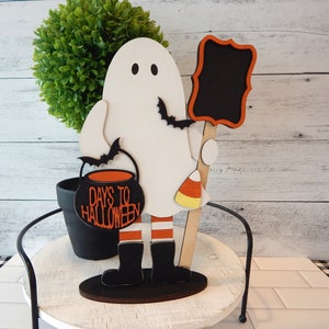 May include: A white ghost figurine holding a black cauldron with the text "Days to Halloween" and a black chalkboard sign. The ghost is wearing orange and white striped stockings and black boots. The figurine is standing on a wooden base.