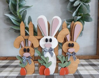 May include: Three wooden bunny figurines stand in front of a wooden picket fence. The bunnies are painted white, brown, and brown with black and white checkered bow ties. Each bunny holds a green plant and a carrot.