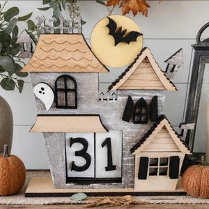 Puede incluir: Un calendario de Halloween de madera con forma de casa embrujada. La casa es gris con detalles de madera, un recorte de fantasma y una silueta de murci&eacute;lago contra una luna amarilla. El calendario muestra el n&uacute;mero 31. Dos calabazas y follaje oto&ntilde;al completan la escena.