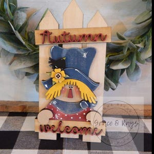 May include: Wooden autumn-themed decoration featuring a scarecrow figure behind a white picket fence. The word "Autumn" is at the top, and "welcome" at the bottom. The scarecrow has a blue hat, yellow hair, and a red and white polka-dot shirt.