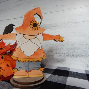 May include: A wooden cutout of a scarecrow with a brown hat, orange shirt, blue jeans, and a white beard. The scarecrow is holding a small black bird in its left hand. The scarecrow is standing on a wooden base.