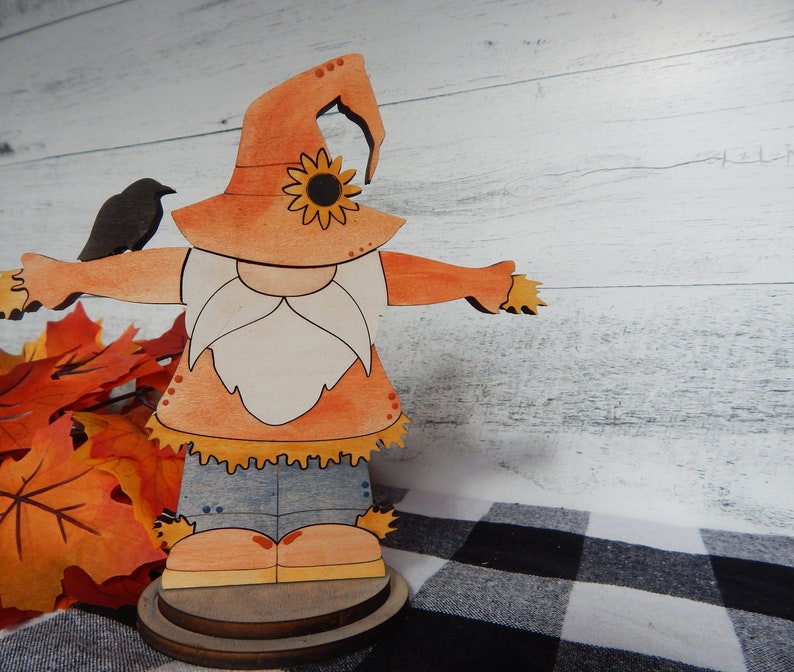 May include: A wooden cutout of a scarecrow with a brown hat, orange shirt, blue jeans, and a white beard. The scarecrow is holding a small black bird in its left hand. The scarecrow is standing on a wooden base.